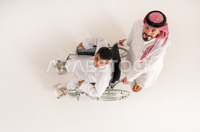 Looking at the camera with gestures of happiness and pleasure, family support and encouragement for people with disabilities, improving the lives of people with motor disabilities to move, vertical top portrait of a Saudi Arabian Gulf man wearing traditional dress helping his son with special needs, white background