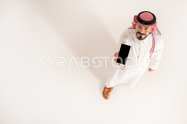 استخدام الأجهزة التقنية الحديثة في الحياة اليومية, النظر إلى الكاميرا بإيماءات السعادة والسرور, بورتريه علوي عامودي لرجل عربي خليجي سعودي مبتسم يرتدي الثوب التقليدي والشماغ يمسك بيده الهاتف المحمول, عرض شاشة سوداء فارغة عبر الجوال, خلفية بيضاء