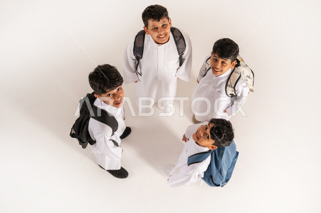 النظر إلى الكاميرا بإيماءات السعادة والسرور, الاستعداد للعام الدراسي الجديد، موسم العودة للمدارس في السعودية, بورتريه علوي عامودي لأولاد عربيين خليجيين سعوديين يرتدون الثوب التقليدي يقفون باستقامة ويحملون الحقائب على ظهرهم, خلفية بيضاء