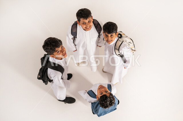 النظر إلى الكاميرا بإيماءات السعادة والسرور, الاستعداد للعام الدراسي الجديد، موسم العودة للمدارس في السعودية, بورتريه علوي عامودي لأولاد عربيين خليجيين سعوديين يرتدون الثوب التقليدي يقفون باستقامة ويحملون الحقائب على ظهرهم, خلفية بيضاء