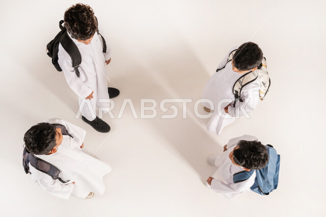 الاستعداد للعام الدراسي الجديد، موسم العودة للمدارس في السعودية, بورتريه علوي عامودي لأولاد عربيين خليجيين سعوديين يرتدون الثوب التقليدي يقفون باستقامة ويحملون الحقائب على ظهرهم, خلفية بيضاء
