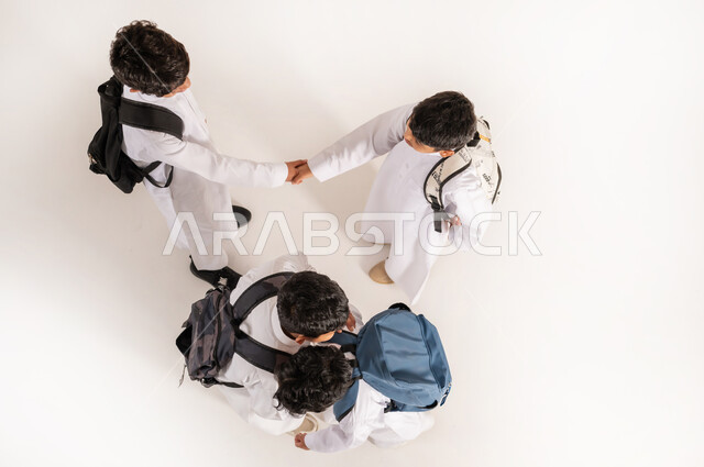 التصافح وتماسك الأيدي بتعابير الاتفاق, الاستعداد للعام الدراسي الجديد، موسم العودة للمدارس في السعودية, بورتريه علوي عامودي لأولاد عربيين خليجيين سعوديين يرتدون الثوب التقليدي يقفون باستقامة ويحملون الحقائب على ظهرهم, خلفية بيضاء