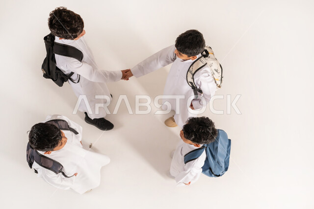 التصافح وتماسك الأيدي بتعابير الاتفاق, الاستعداد للعام الدراسي الجديد، موسم العودة للمدارس في السعودية, بورتريه علوي عامودي لأولاد عربيين خليجيين سعوديين يرتدون الثوب التقليدي يقفون باستقامة ويحملون الحقائب على ظهرهم, خلفية بيضاء