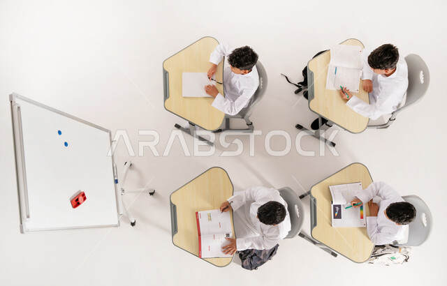 Concept of diligence and excellence, modern learning methods, back to school season, goals of education in the Kingdom of Saudi Arabia, vertical top portrait of Saudi Arabian Gulf Arab students wearing traditional dress sitting on desks in the classroom with school books in front of them, white background