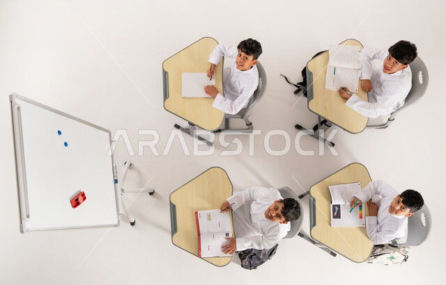 النظر إلى الكاميرا بإيماءات السعادة والسرور, مفهوم المشاركة الفعالة, أساليب التعلم الحديثة، موسم العودة إلى المدارس, بورتريه علوي عامودي لطلاب عربيين خليجيين سعوديين يرتدون الثوب التقليدي يجلسون على المقاعد داخل الصف وأمامهم الكتب المدرسية, خلفية بيضاء