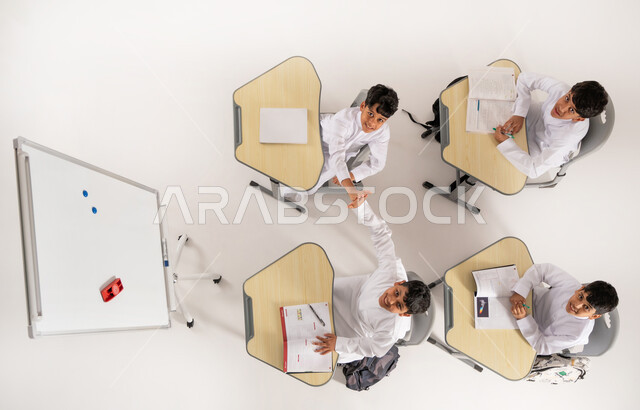 Back to school season, holding hands with expressions of success and agreement, looking at camera with gestures of happiness, concept of active participation, vertical overhead portrait of Saudi Arabian Gulf students wearing traditional dress sitting on desks in classroom with school books in front of them, white background