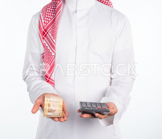 بورتريه لرجل عربي خليجي سعودي، يحمل في يده عملة ورقية والة حاسبة في اليد الاخرى، العملات الورقية للريال السعودية، استخدام الالة الحاسبة لاجراء العمليات الحسابية، ادارة الحسابات