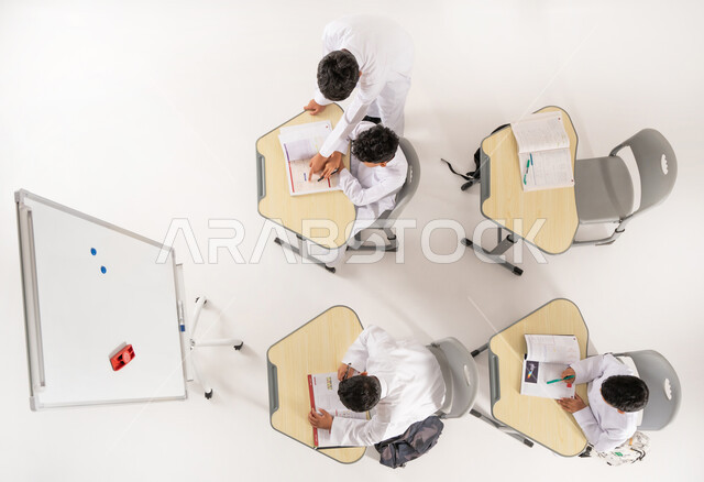 موسم العودة إلى المدارس, أهداف التربية والتعليم في المملكة العربية السعودية, أساليب التعلم الحديثة, مفهوم المشاركة الفعالة، بورتريه علوي عامودي لطلاب عربيين خليجيين سعوديين يرتدون الثوب التقليدي يجلسون على المقاعد داخل الصف وأمامهم الكتب المدرسية, خلفية بيضاء