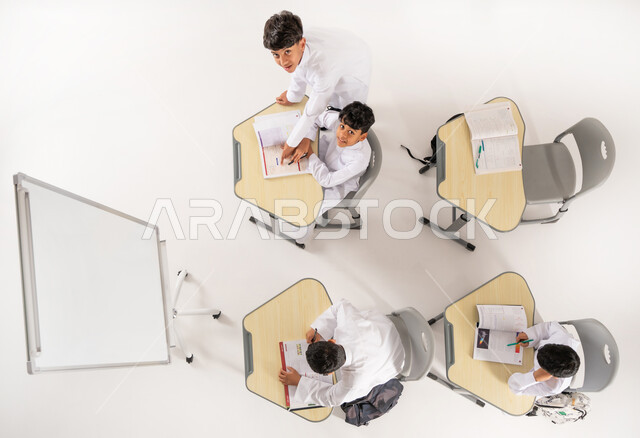 النظر إلى الكاميرا بإيماءات السعادة والسرور, مفهوم المشاركة الفعالة, أساليب التعلم الحديثة، موسم العودة إلى المدارس, بورتريه علوي عامودي لطلاب عربيين خليجيين سعوديين يرتدون الثوب التقليدي يجلسون على المقاعد داخل الصف وأمامهم الكتب المدرسية, خلفية بيضاء