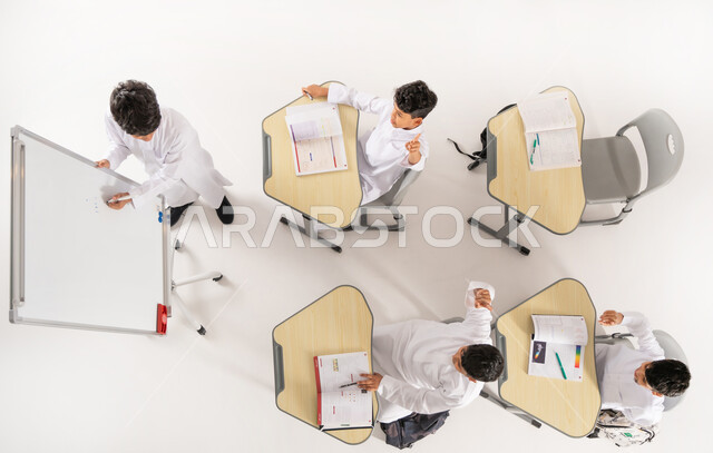 مفهوم المشاركة الفعالة, أساليب التعلم الحديثة، موسم العودة إلى المدارس, أهداف التربية والتعليم في المملكة العربية السعودية، بورتريه علوي عامودي لطلاب عربيين خليجيين سعوديين يرتدون الثوب التقليدي يجلسون على المقاعد داخل الصف وأمامهم الكتب المدرسية, خلفية بيضاء