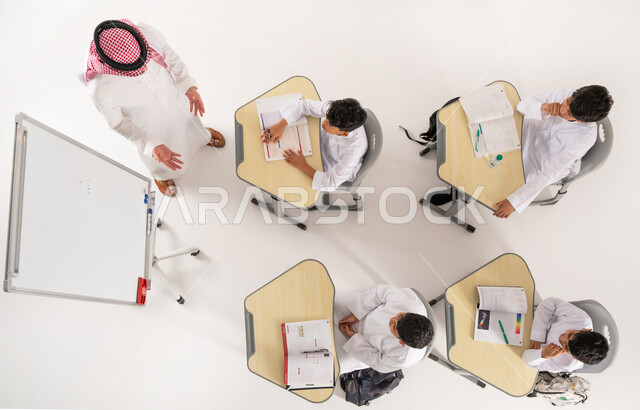 أهداف التربية والتعليم في المملكة العربية السعودية, مفهوم المشاركة الفعالة, أساليب التعلم الحديثة، موسم العودة إلى المدارس، بورتريه علوي عامودي لمعلم عربي خليجي سعودي يرتدي الثوب التقليدي والشماغ يقوم بشرح الدرس للطلاب داخل الصف, خلفية بيضاء