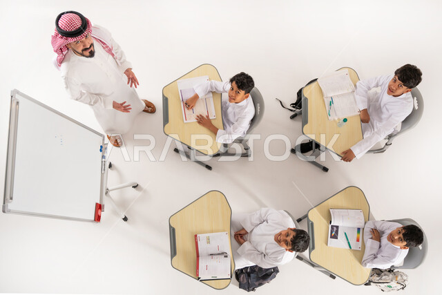 اكتساب المعرفة و الخبرات العلمية، بورتريه علوي عامودي لمعلم عربي خليجي سعودي يقوم بإعطاء الدروس، تلاميذ عرب خليجيون سعوديون يرتدون الثوب السعودي التقليدي يجلسون على الكراسي وأمامهم طاولات الدراسة، المثابرة على الدراسة لتحقيق النجاح، خلفية بيضاء