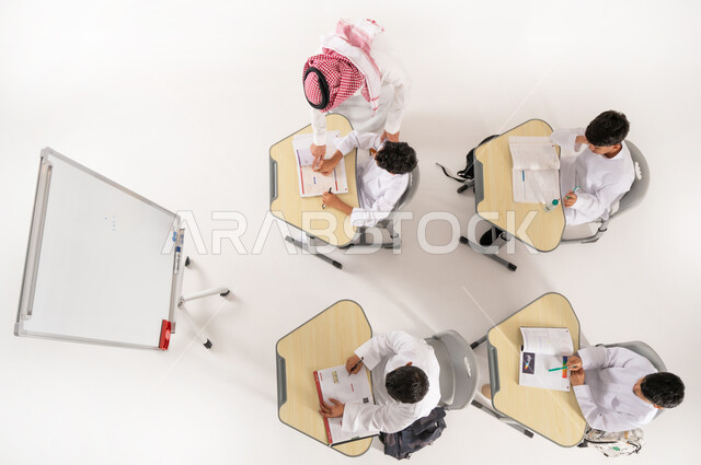 دور المدارس في تحقيق التطور الشخصي، مفهوم الاهتمام بالعلم والتعليم، تلاميذ عرب خليجيون سعوديون يرتدون الثوب التقليدي جلسون في الصف، القيام بكتابة الواجبات المدرسية، بورتريه علوي عامودي لمعلم عربي خليجي سعودي يقوم بمساعدة الطلاب بعمل نشاطات جماعية، خلفية بيضاء