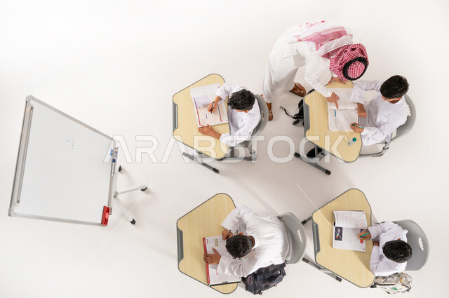 مفهوم الاهتمام بالعلم والتعليم، تلاميذ عرب خليجيون سعوديون يرتدون الثوب التقليدي جلسون في الصف، القيام بكتابة الواجبات المدرسية، بورتريه علوي عامودي لمعلم عربي خليجي سعودي يقوم بمساعدة الطلاب بعمل نشاطات جماعية، دور المدارس في تحقيق التطور الشخصي، خلفية بيضاء