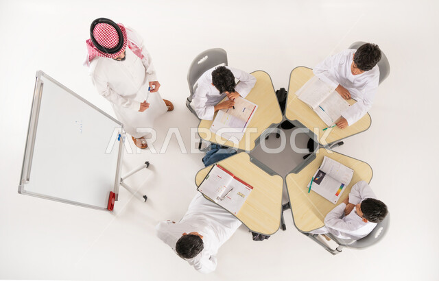 القيام بكتابة الواجبات المدرسية، بورتريه علوي عامودي لمعلم عربي خليجي سعودي يقوم بمساعدة الطلاب بعمل نشاطات جماعية، دور المدارس في تحقيق التطور الشخصي، مفهوم الاهتمام بالعلم والتعليم، تلاميذ عرب خليجيون سعوديون يرتدون الثوب التقليدي جلسون في الصف، خلفية بيضاء