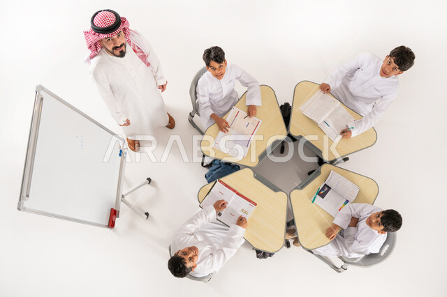 النظر الى الكاميرا بايماءات الحماس والسعادة، بورتريه علوي عامودي لمعلم عربي خليجي سعودي يقوم بإعطاء الدروس، تلاميذ عرب خليجيون سعوديون يرتدون الثوب السعودي التقليدي يجلسون بشكل مجموعة، المثابرة على الدراسة لتحقيق النجاح، الجلسات والحصص التعليمية، خلفية بيضاء