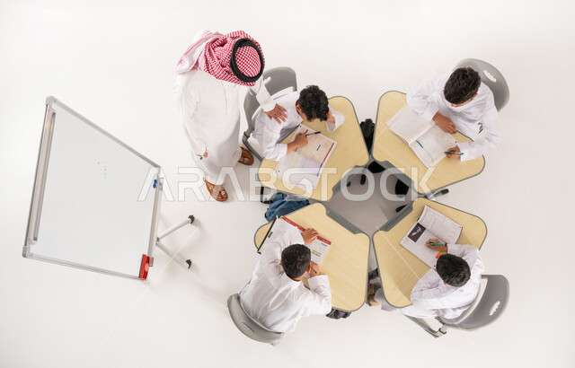 دور المدارس في تحقيق التطور الشخصي، مفهوم الاهتمام بالعلم والتعليم، تلاميذ عرب خليجيون سعوديون يرتدون الثوب التقليدي جلسون في الصف، القيام بكتابة الواجبات المدرسية، بورتريه علوي عامودي لمعلم عربي خليجي سعودي يقوم بمساعدة الطلاب بعمل نشاطات جماعية، خلفية بيضاء
