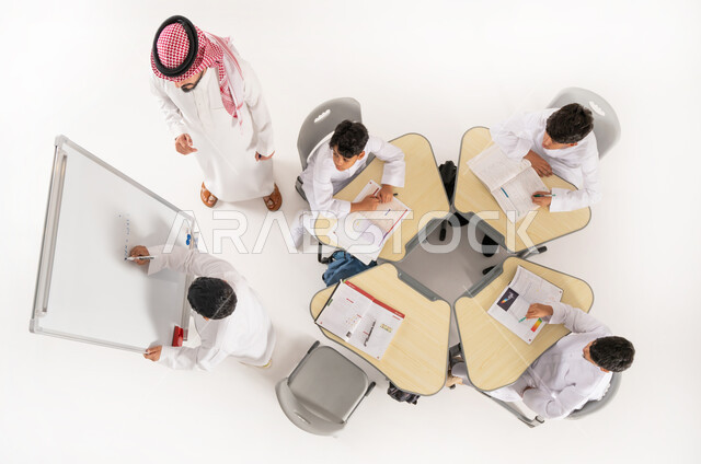 الاهتمام بالعملية التعليمية، انشطة جماعية لتعليم الطلاب في المدارس، تلاميذ عرب خليجيون سعوديون يرتدون الثوب السعودي التقليدي يجلسون على الكراسي وأمامهم طاولات الدراسة، بورتريه علوي عامودي لمعلم عربي خليجي سعودي يقوم بإعطاء الدروس، طالب يشرح فكرة ما، خلفية بيضاء