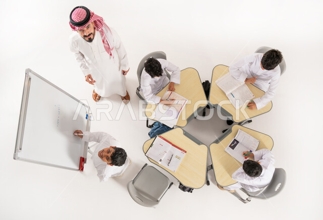 انشطة جماعية لتعليم الطلاب في المدارس، تلاميذ عرب خليجيون سعوديون يرتدون الثوب السعودي التقليدي يجلسون على الكراسي وأمامهم طاولات الدراسة، بورتريه علوي عامودي لمعلم عربي خليجي سعودي يقوم بإعطاء الدروس، طالب يشرح فكرةما، الاهتمام بالعملية التعليمية، خلفية بيضاء