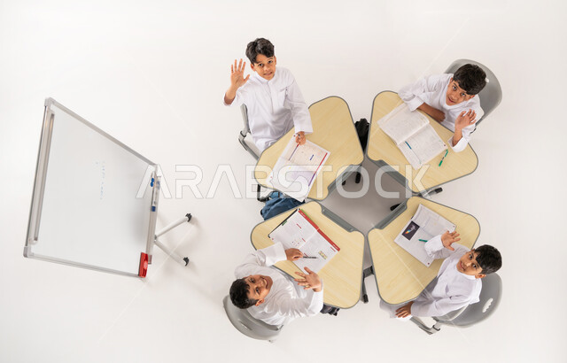 النظر الى الكاميرا بتعابير الفرح والسرور، الاهتمام بالعلم والتعلم، مفهوم الاجتهاد و طلب التفوق، التعليم الدراسي في المملكة السعودية، بورتريه علوي عامودي لتلاميذ عرب خليجيون سعوديون يرتدون الثوب السعودي التقليدي يرفعون ايديهم بإيماءات الحماس، خلفية بيضاء