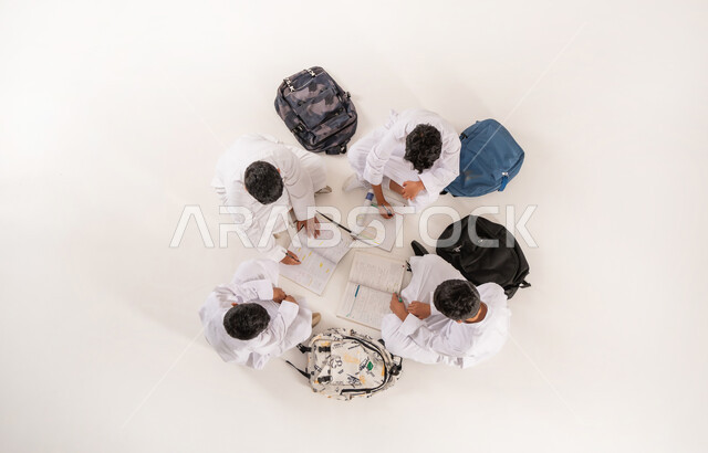Expressions of concentration and engagement in lessons, developing creative skills, vertical top portrait of a group of Saudi Arabian Gulf boys wearing traditional dress sitting on the floor, enjoying group recreational activities, writing homework with friends, white background