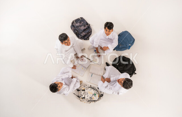 كتابة الواجبات المنزلية مع الأصدقاء، النظر إلى الكاميرا بايماءات السرور والسعادة، تنمية المهارات الابداعية، بورتريه علوي عامودي لمجموعة أولاد عربيين خليجيين سعوديين يرتدون الثوب التقليدي يجلسون على الأرض, الاستمتاع بالأنشطة الترفيهية الجماعية, خلفية بيضاء