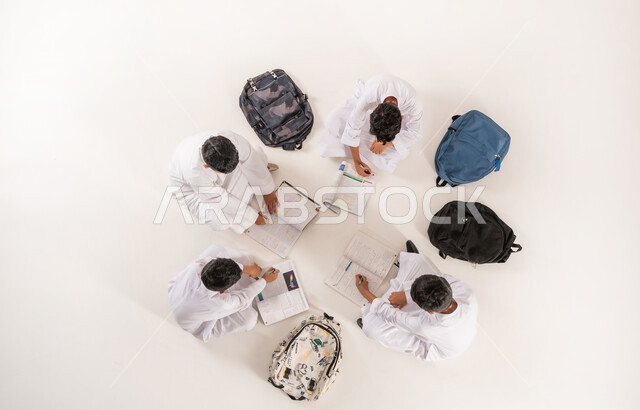 الاستمتاع بالأنشطة الترفيهية الجماعية, كتابة الواجبات المنزلية مع الأصدقاء، تعابير التركيز والاندماج  في الدروس، تنمية المهارات الابداعية، بورتريه علوي عامودي لمجموعة أولاد عربيين خليجيين سعوديين يرتدون الثوب التقليدي يجلسون على الأرض, خلفية بيضاء