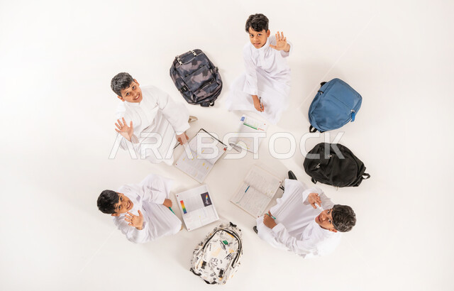 Developing creative skills, vertical overhead portrait of a group of Saudi Arabian Gulf boys wearing traditional dress sitting on the floor, enjoying group leisure activities, writing homework with friends, looking at the camera with gestures of happiness and pleasure, white background