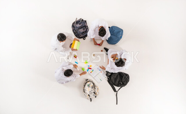 قضاء وقت ممتع مع الرفاق في المدرسة، ممارسة الهوايات المفضلة مع الآخرين، استخدام الوسائل الحديثة في الدراسة, مشاركة الأصدقاء في التعلم و اللعب, بورتريه علوي عامودي لمجموعة أولاد عربيين خليجيين سعوديين يرتدون الثوب التقليدي  يجلسون على الأرض, خلفية بيضاء
