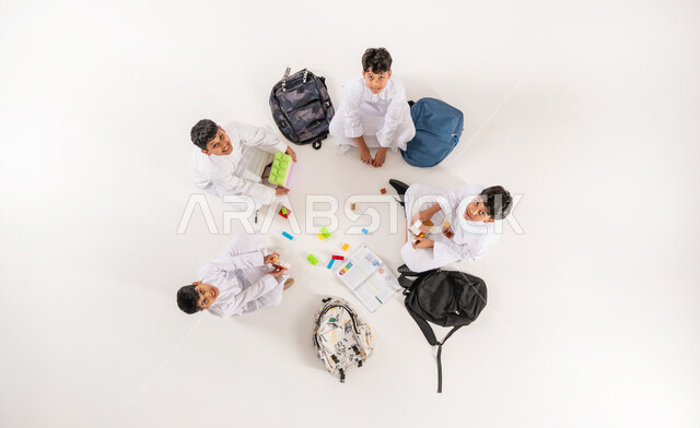 ممارسة الهوايات المفضلة مع الآخرين، النظر إلى الكاميرا بايماءات السعادة والسرور، مشاركة الأصدقاء في التعلم واللعب, بورتريه علوي عامودي لمجموعة أولاد عربيين خليجيين سعوديين يرتدون الثوب التقليدي  يجلسون على الأرض, قضاء وقت ممتع مع الرفاق في المدرسة، خلفية بيضاء