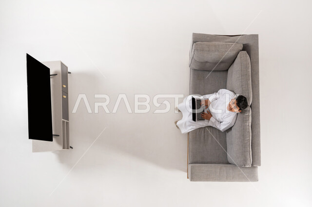 التعلم عن بعد عبر الأجهزة التقنية الحديثة, بورتريه علوي عامودي لولد عربي خليجي سعودي يرتدي الثوب التقليدي  يجلس على الأريكة في غرفة المعيشة ويستخدم جهاز الحاسب المحمول، النظر إلى الكاميرا بايماءات السعادة، عرض شاشة سوداء فارغة عبر التلفاز والحاسب, خلفية بيضاء