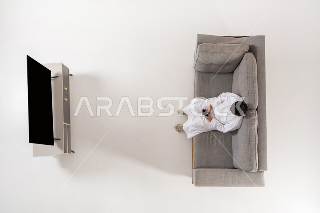 قضاء وقت الفراغ باستخدام التقنيات الحديثة, بورتريه علوي عامودي لولد عربي خليجي سعودي يرتدي الثوب التقليدي يجلس على الأريكة في غرفة المعيشة يعمل على جهاز الهاتف المحمول بايماءات الاندماج, لعب ألعاب الفيديو المسلية والممتعة, عرض شاشة سوداء فارغة، خلفية بيضاء