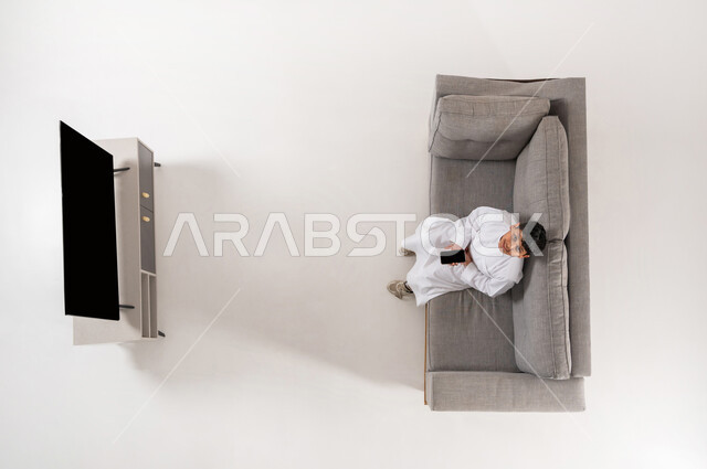 ألعاب الفيديو المسلية, بورتريه علوي عامودي لولد عربي خليجي سعودي يرتدي الثوب التقليدي يجلس على الأريكة في غرفة المعيشة يعمل على جهاز الهاتف المحمول، النظر إلى الكاميرا بإيماءات السعادة, عرض شاشة سوداء فارغة، الترفيه باستخدام التكنولوجيا الحديثة, خلفية بيضاء