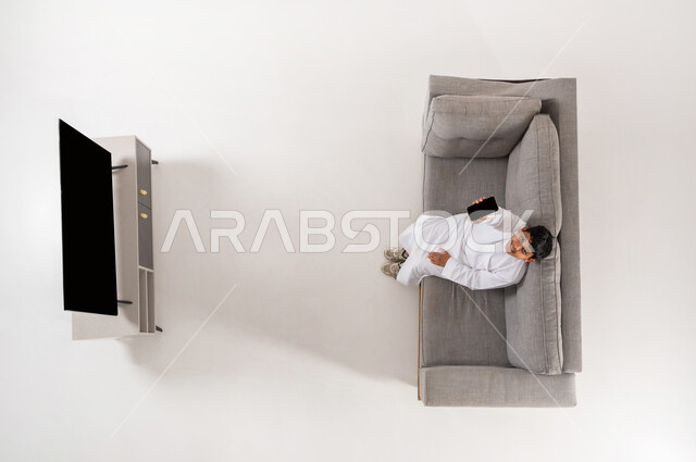 الترفيه باستخدام التكنولوجيا الحديثة, ألعاب الفيديو المسلية, بورتريه علوي عامودي لولد عربي خليجي سعودي يرتدي الثوب التقليدي يجلس على الأريكة في غرفة المعيشة يعمل على جهاز الهاتف المحمول، النظر إلى الكاميرا بإيماءات السعادة, عرض شاشة سوداء فارغة، خلفية بيضاء