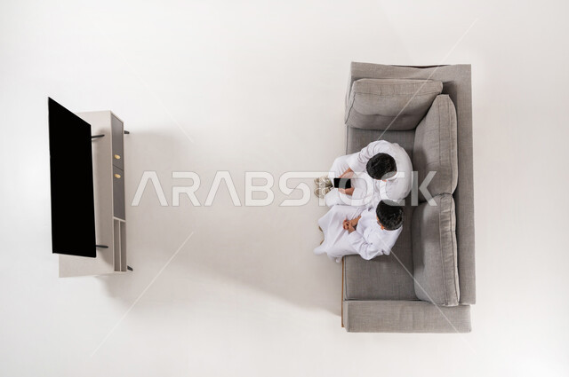 Gestures of interaction and integration, sharing electronic entertainment activities with friends, gaining free time by practicing hobbies, vertical top portrait of two Saudi Arabian Gulf boys wearing traditional dress sitting on sofa in living room, black blank screen display, white background