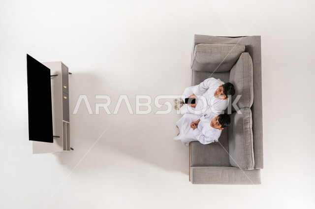 عرض شاشة سوداء فارغة، النظر إلى الكاميرا بايماءات الفرح، مشاركة الأصدقاء بالأنشطة الترفيهية الالكترونية, بورتريه علوي عامودي لولدان عربيان خليجييان سعوديان يرتديان الثوب التقليدي يجلسان على الأريكة في غرفة المعيشة يعملان على الهاتف المحمول، خلفية بيضاء