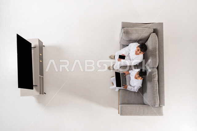 النظر إلى الكاميرا بايماءات الفرح، مشاركة الأصدقاء بالأنشطة الترفيهية الالكترونية, بورتريه علوي عامودي لولدان عربيان خليجييان سعوديان يرتديان الثوب التقليدي يجلسان على الأريكة في غرفة المعيشة يعملان على الأجهزة اللوحية، عرض شاشة سوداء فارغة، خلفية بيضاء