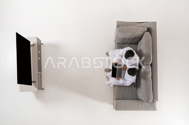 مشاركة الأصدقاء بالأنشطة الترفيهية الالكترونية، اكتساب وقت الفراغ بممارسة الهوايات, بورتريه علوي عامودي لولدان عربيان خليجييان سعوديان يرتديان الثوب التقليدي يجلسان على الأريكة في غرفة المعيشة, عرض شاشة سوداء فارغة، إيماءات التفاعل و الاندماج , خلفية بيضاء