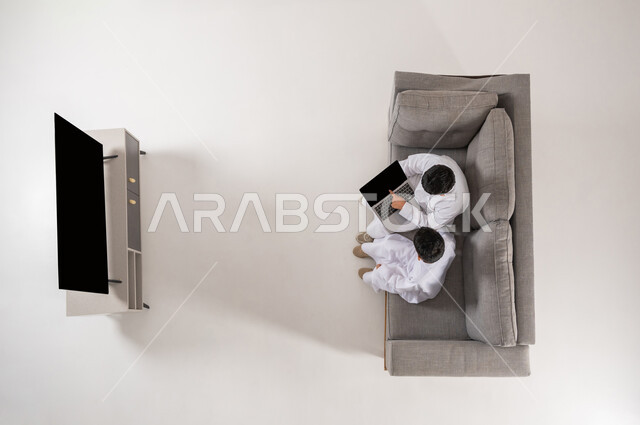 إيماءات التفاعل و الاندماج, مشاركة الأصدقاء بالأنشطة الترفيهية الالكترونية في أوقات الفراغ, بورتريه علوي عامودي لولدان عربيان خليجييان سعوديان يرتديان الثوب التقليدي يجلسان على الأريكة في غرفة المعيشة, الإشارة بإصبع السبابة إلى شاشة سوداء فارغة، خلفية بيضاء