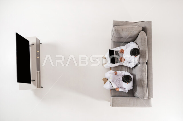 القيام بالأنشطة الترفيهية, مشاركة الأصدقاء في هواية المطالعة, بورتريه علوي عامودي لولدان عربيان خليجييان سعوديان يرتديان الثوب التقليدي يجلسان على الأريكة في غرفة المعيشة , تنمية القراءة عند الأولاد باستخدام التقنيات الحديثة, عرض شاشة سوداء فارغة, خلفية بيضاء