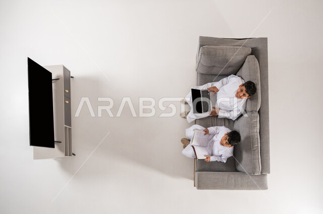 تنمية القراءة عند الأولاد باستخدام التقنيات الحديثة, عرض شاشة سوداء فارغة, النظر إلى الكاميرا بايماءات الفرح، القيام بالأنشطة الترفيهية, بورتريه علوي عامودي لولدان عربيان خليجييان سعوديان يرتديان الثوب التقليدي يجلسان على الأريكة في غرفة المعيشة, خلفية بيضاء