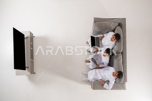 النظر الى الكاميرا بايماءات السعادة, عرض شاشة سوداء فارغة، ممارسة هواية المطالعة عن طريق جهاز اللابتوب والكتب, بورتريه علوي عامودي لثلاثة أولاد عربيين سعوديين خليجييين يرتدون الثوب التقليدي يجلسون على الأريكة في غرفة المعيشة يقومون بقراءة القصص, خلفية بيضاء