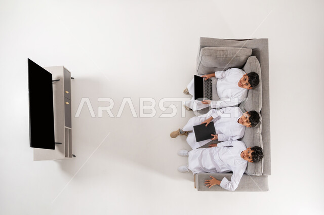 المشاركة في قضاء الأوقات المسلية, بورتريه علوي عامودي لثلاثة أولاد عربيين سعوديين خليجيين يرتدون الثوب التقليدي يجلسون على الأريكة في غرفة المعيشة، عرض شاشة سوداء فارغة، استخدام الأجهزة الحديثة مع الأصدقاء, النظر الى الكاميرا بايماءات السرور، خلفية بيضاء