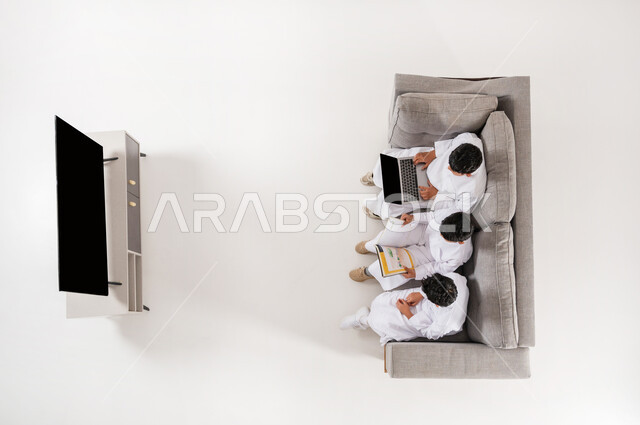 ممارسة هواية المطالعة عن طريق جهاز اللابتوب والكتب, بورتريه علوي عامودي لثلاثة أولاد عربيين سعوديين خليجييين يرتدون الثوب التقليدي يجلسون على الأريكة في غرفة المعيشة يقومون بقراءة القصص, الإشارة باليد و الاندماج و المشاركة, عرض شاشة سوداء فارغة، خلفية بيضاء