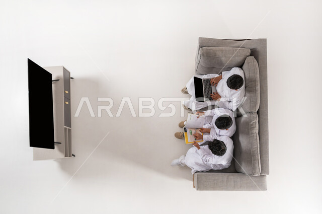 الإشارة باليد و الاندماج و المشاركة, عرض شاشة سوداء فارغة، ممارسة هواية المطالعة عن طريق الجهاز اللوحي والكتب, بورتريه علوي عامودي لثلاثة أولاد عربيين سعوديين خليجييين يرتدون الثوب التقليدي يجلسون على الأريكة في غرفة المعيشة يقومون بقراءة القصص, خلفية بيضاء