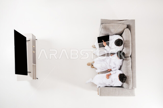 المشاركة في قضاء الأوقات المسلية, بورتريه علوي عامودي لثلاثة أولاد عربيين سعوديين خليجيين يرتدون الثوب التقليدي يجلسون على الأريكة في غرفة المعيشة، عرض شاشة سوداء فارغة، الإشارة على جهاز التابلت،استخدام الأجهزة الحديثة في اللعب والتعلم مع الأصدقاء, خلفية بيضاء