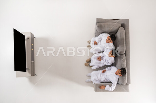 الاستمتاع بقضاء الوقت مع الأصدقاء , بورتريه علوي عامودي لثلاثة أولاد عربيين سعوديين خليجيين يرتدون الثوب التقليدي يجلسون على الأريكة في غرفة المعيشة بتعابير البشاشة، عرض شاشة سوداء فارغة عبر التلفاز، النظر إلى الكاميرا بإيماءات السعادة و السرور, خلفية بيضاء