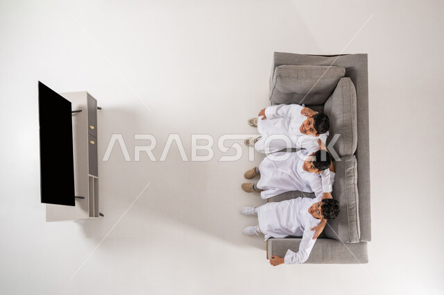 النظر إلى الكاميرا بإيماءات السعادة و السرور, الاستمتاع بقضاء الوقت مع الأصدقاء , بورتريه علوي عامودي لثلاثة أولاد عربيين سعوديين خليجيين يرتدون الثوب التقليدي يجلسون على الأريكة في غرفة المعيشة بتعابير البشاشة، عرض شاشة سوداء فارغة عبر التلفاز، خلفية بيضاء