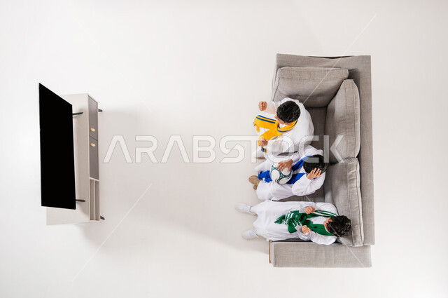 مشاركة الأصدقاء الأوقات الممتعة, بورتريه علوي عامودي لثلاثة أولاد عربيين سعوديين خليجييين يرتدون الثوب التقليدي  يجلسون على الأريكة في غرفة المعيشة, متابعة مباراة كأس العالم, إيماءات التشجيع و الحماس الفوز الخسارة, عرض شاشة سوداء فارغة عبر التلفاز, خلفية بيضاء