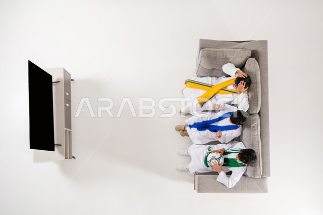 متابعة مباراة كأس العالم, إيماءات التشجيع و الحماس الفوز الخسارة, عرض شاشة سوداء فارغة عبر التلفاز, مشاركة الأصدقاء الأوقات الممتعة, بورتريه علوي عامودي لثلاثة أولاد عربيين سعوديين خليجييين يرتدون الثوب التقليدي  يجلسون على الأريكة في غرفة المعيشة, خلفية بيضاء